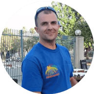 Smiling man in blue t-shirt, eMachineShop customer, against fence background.