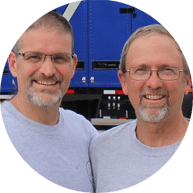Two smiling men in gray shirts, a clients of eMachineShop, standing close together with a blue truck in the background.